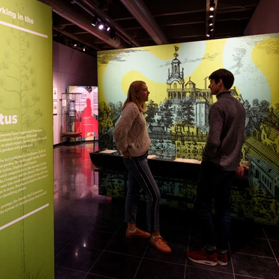 Two young visitors conversing in front of a large illustrated wall showing Leiden’s historic botanical garden and university buildings, with nearby panels on botanical research.