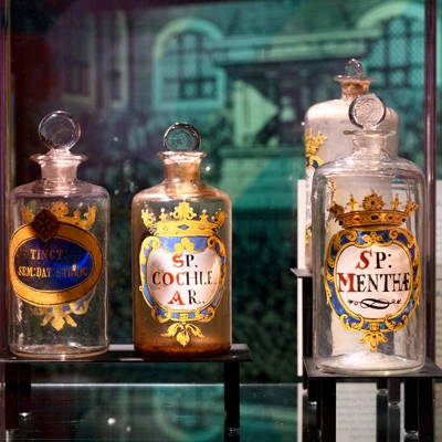 Close-up of four ornate apothecary jars labeled in Latin, each adorned with a decorative crest, arranged inside a glass display against a clean backdrop.