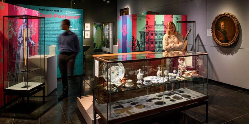 Woman smiling as she observes a glass case displaying fine porcelain tableware, set within a gallery of vivid wall illustrations and a historic portrait hanging in the background.