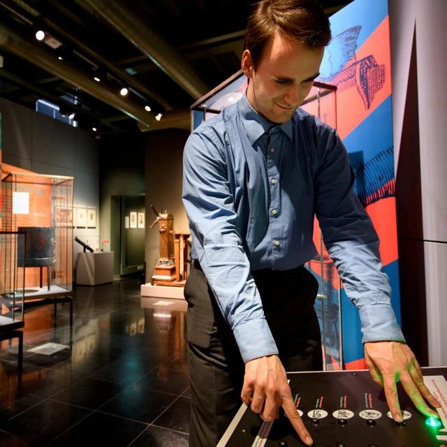 A man in a blue shirt using a tactile interactive station, with buttons and dials, in a gallery filled with historic scientific tools and instructional displays.