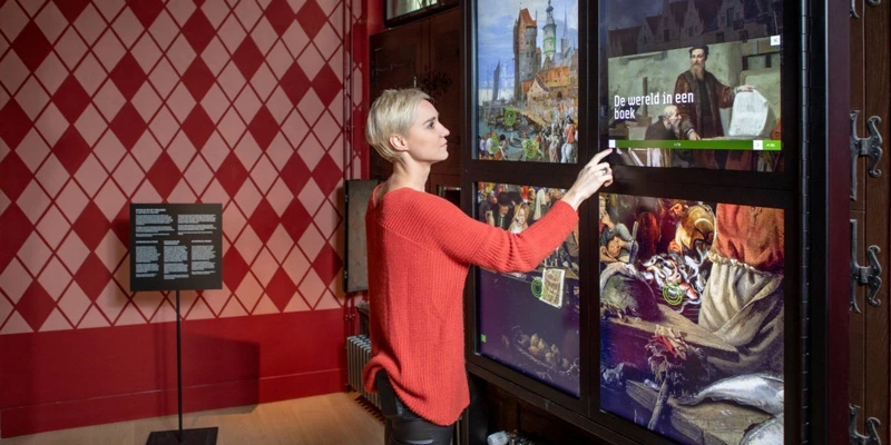 Female visitor touching a vertical touchscreen display embedded in a large cabinet structure, revealing layered stories through artworks and animated content.