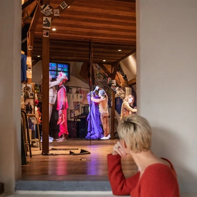 Distant view of an immersive theatrical installation, with a woman seated in the foreground watching life-sized projections of actors in costume performing inside a recreated artist’s studio.