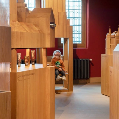 Child seated among oversized wooden architectural models, gazing up in awe at a screen embedded in the wooden structure above.