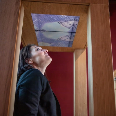 Woman looking up inside a vertical wooden structure with a digital ceiling screen displaying a kaleidoscopic architectural animation.