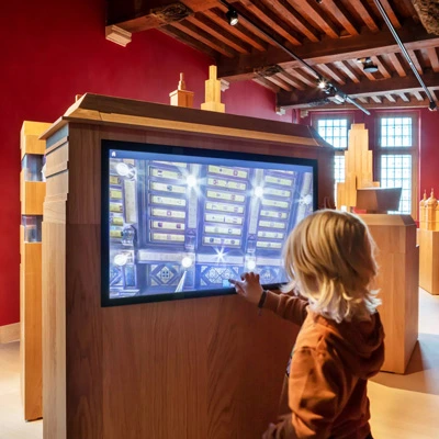 Child interacting with a large touchscreen display embedded in a wooden exhibit structure, surrounded by architectural models and warm lighting.