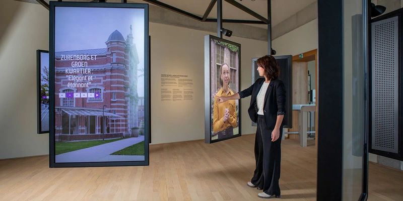 Woman exploring a room lined with life-size vertical screens, each presenting cultural stories and visuals from different neighborhoods of Antwerp.