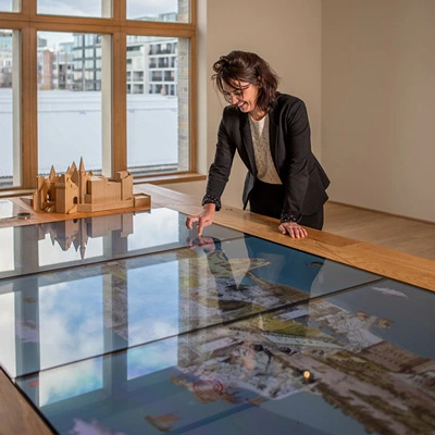 Woman using a large horizontal touchscreen map alongside a wooden scale model of Antwerp, in a sunlit room with tall windows.