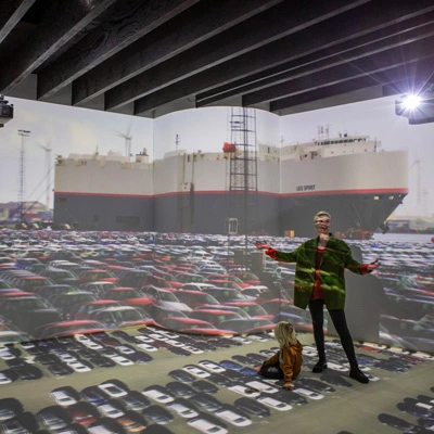 Woman and child inside an immersive panoramic projection room with floor-to-ceiling visuals of a car terminal and cargo ship.