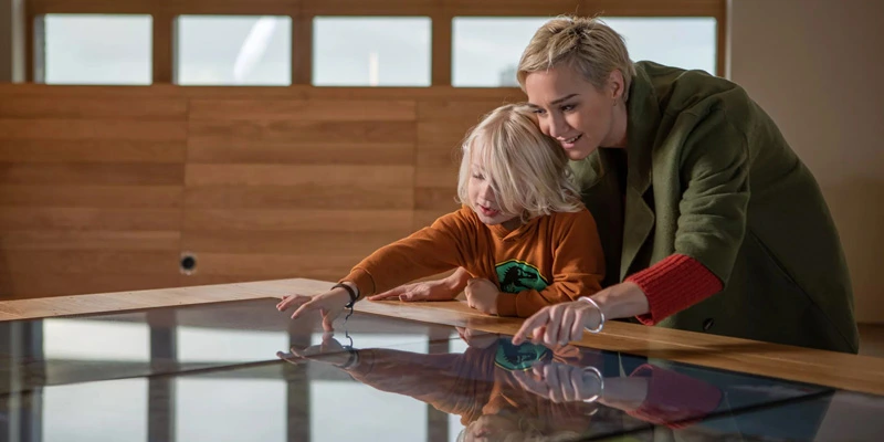 Woman and child engaging with a large interactive map table; both smiling and pointing to different elements on the digital surface in a warmly lit room.