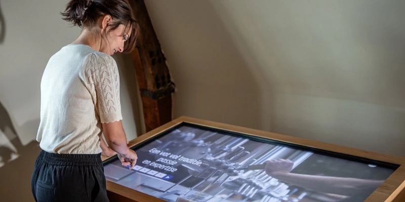 Woman interacting with a horizontal touchscreen exhibit focused on craftsmanship and local industry, featuring large visuals and text in a softly lit space.