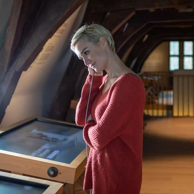Woman standing in a dimly lit attic space, listening to an audio track through a handset while interacting with a touchscreen display embedded in a wooden podium.
