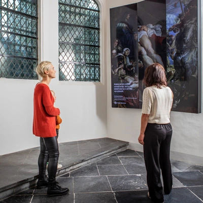 Two women and a child observing a large vertical digital display showing a high-resolution reproduction of a classical painting, inside a bright gallery space with tall windows.