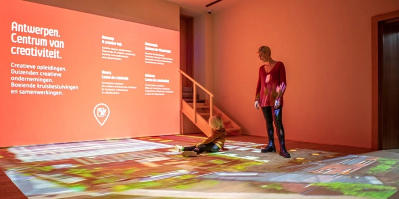 A child sits on the floor while a woman stands nearby, both immersed in an interactive installation where colorful visuals and texts about Antwerp’s creative industry are projected onto the floor and wall.