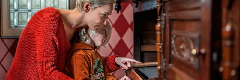 Parent and child closely examining the drawers of a historic wooden cabinet in a red-patterned room at Het Steen, discovering tactile and visual elements of Antwerp’s past.