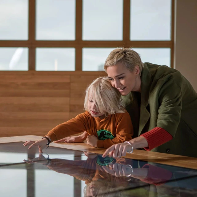 Woman and child engaging with a large interactive map table; both smiling and pointing to different elements on the digital surface in a warmly lit room.