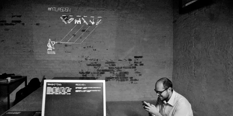 Black-and-white photograph of an art installation room. An interactive display mapping a satellite dish to deep space probes is projected on a brick wall. In the foreground, a computer monitor shows data logs next to a seated person using their smartphone.