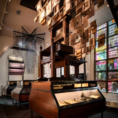 Display on bookmaking, featuring a historical printing press surrounded by books and miniature figures, with suspended printed pages above and colorful stained glass at the back.