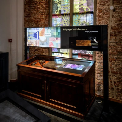 Interactive station with antique tools and a touchscreen, set against a stained glass window featuring collage-style banknote imagery and historical references.