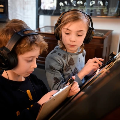 Two children wearing headphones and holding clipboards, engaging with an interactive exhibit on printing techniques and history.