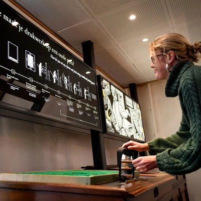 Woman engaging with a hands-on printing exhibit, pulling a lever on a wooden structure below a screen that explains how to set up and use a historical press.