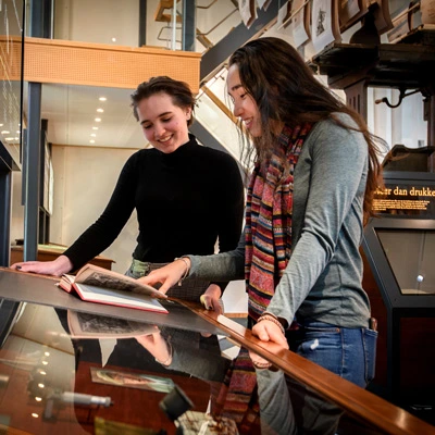 Two young women reading and discussing a book displayed on a glass-topped museum table, surrounded by other printing-related displays and exhibition graphics.