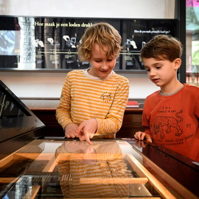 Two boys interacting with an illuminated display table, one pointing to a printing-related exhibit beneath the glass, with educational diagrams about lead type in the background.