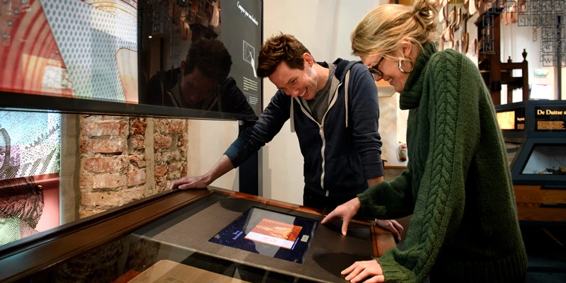 Man and woman smiling as they engage with a digital screen embedded in a display table, surrounded by colorful exhibition elements and brick walls.