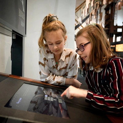 Two young girls interacting with a touchscreen display in the museum, one pointing at the interface while the other attentively observes, with printing equipment visible in the background.