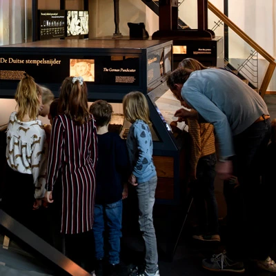 Group of children gathered around a display case labeled “De Duitse stempelsnijder” (“The German Punchcutter”), observing and discussing its contents, with an adult guiding them.