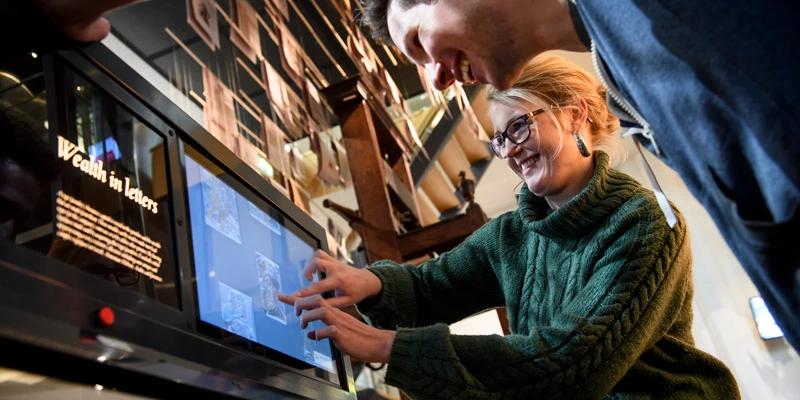 Close-up of two adults smiling and using an interactive digital screen labeled “Wealth in letters,” with a printing press and suspended sheets visible above.