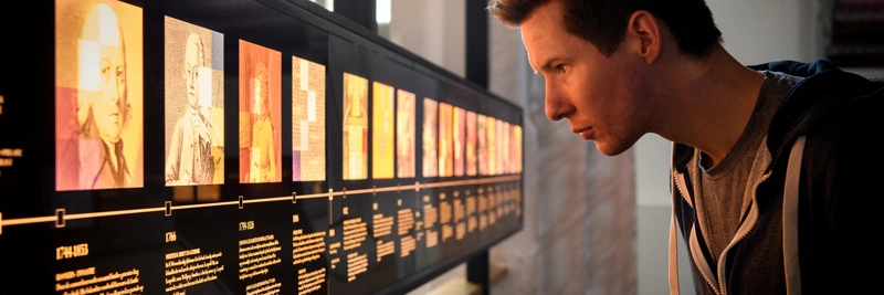 Young man closely inspecting a backlit historical timeline of portraits and key events in the legacy of Joh. Enschedé, a Dutch printer of value documents.