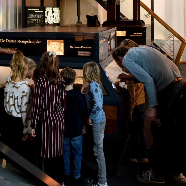 Group of children gathered around a display case labeled “De Duitse stempelsnijder” (“The German Punchcutter”), observing and discussing its contents, with an adult guiding them.