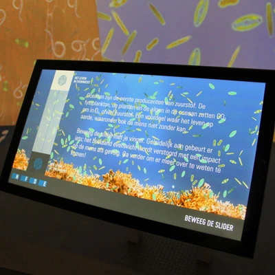 Close-up of a touchscreen showing a Dutch-language interactive explaining how oceans produce oxygen, with visuals of phytoplankton and coral reefs.