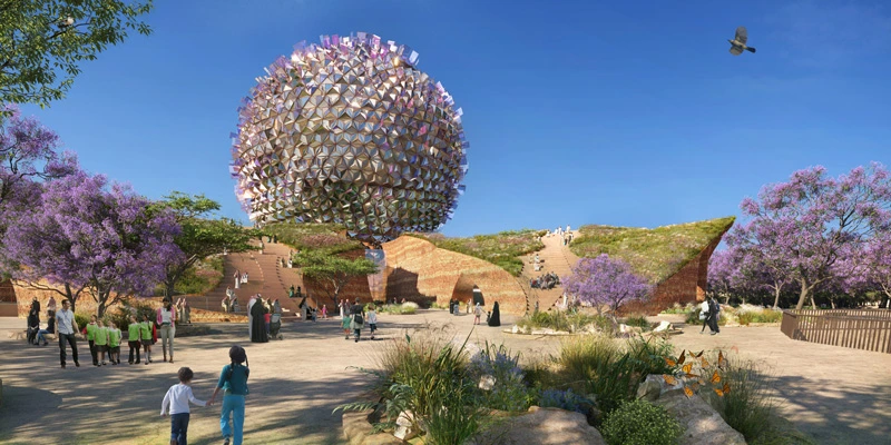 Daytime view of Ilmi Center showing families and school children walking toward the iconic reflective sphere, surrounded by gardens and flowering trees.