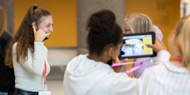 A girl smiles while another student films her using a tablet, capturing a scene for the interactive assignment indoors.