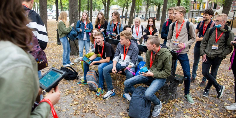 A group of teenage students gathers in a leafy urban area, wearing red lanyards and name badges. Some are seated on a bench while others stand around them, smiling and holding tablets as part of an educational activity.