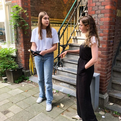 Two girls stand on the sidewalk in front of a staircase, one holding a tablet and the other gesturing while talking, engaged in discussion.