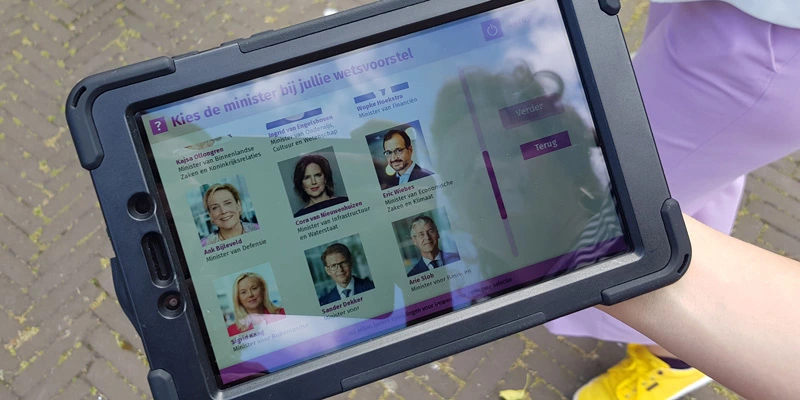 Close-up of a tablet screen showing a digital interface where users select a Dutch government minister as part of the game.