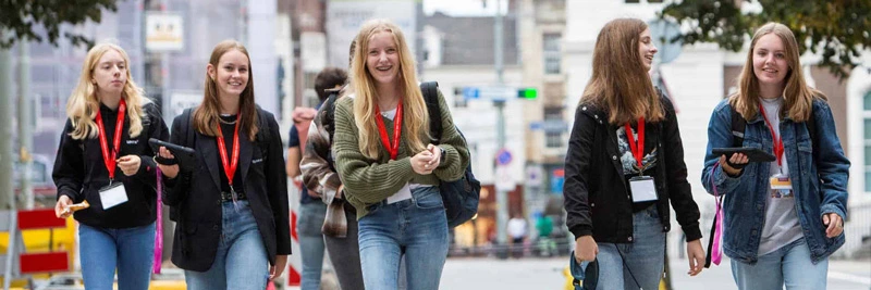 Five teenage girls walk side by side through a city street, smiling and chatting. They wear red lanyards with badges, and some hold tablets as part of an educational activity.