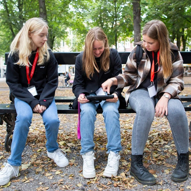 Three girls sit on a park bench, looking at a tablet together, one pointing at the screen as they discuss.