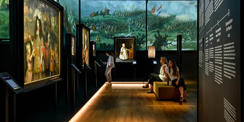 Exhibition room with colonial portraits and a timeline wall. A large painting of a woman accompanied by a white child and a Black child dominates the foreground, while visitors quietly observe other paintings in the surroundings.