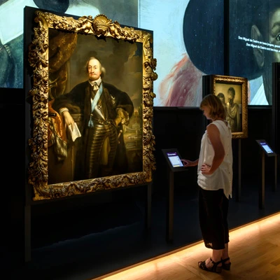 A woman interacts with a touchscreen display in front of a large, ornately framed portrait of Johan Maurits. In the background, projected images and a smaller portraits of two Black men are visible.