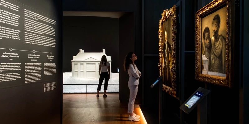 Two women explore a gallery space featuring a detailed white architectural model and framed portraits of Black men. On the left wall, an exhibition timeline in Dutch and English provides historical context.