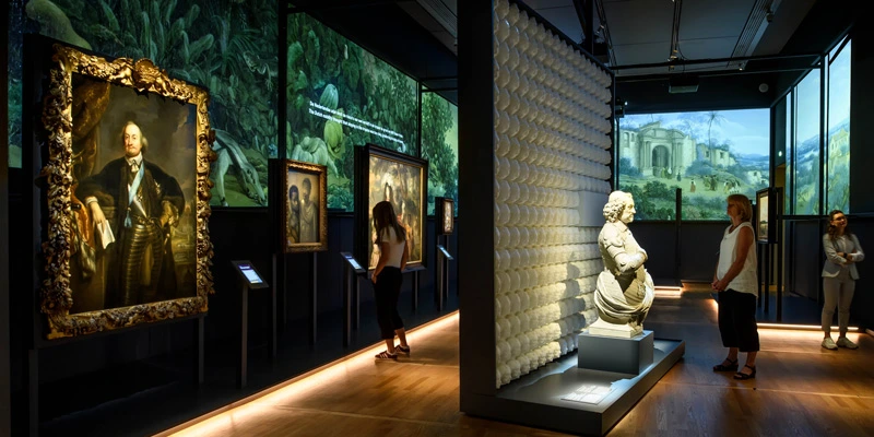 Gallery scene with a bust of Johan Maurits in the center, surrounded by wall-mounted colonial paintings and immersive projections of exotic landscapes. Several visitors explore the room independently.
