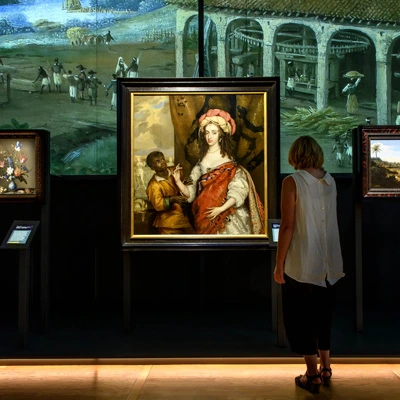 A woman stands closely in front of a large colonial portrait of a white woman accompanied by a Black servant. Projected market scenes and paintings are layered across the back wall.