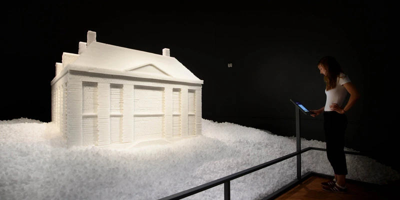 Side view of a woman reading from a touchscreen next to a large white architectural model of a neoclassical building, made of imitation sugar cubes. The installation is surrounded by a base of loose sugar cubes and illuminated in a dark room.