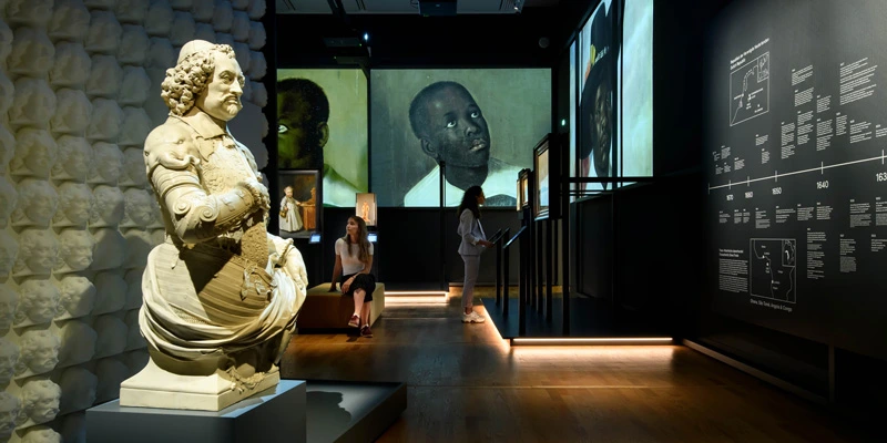 Panoramic view of the exhibition room with a timeline wall, large projected colonial portraits, and a seated visitor. The bust of Johan Maurits appears in the foreground, partially turned.