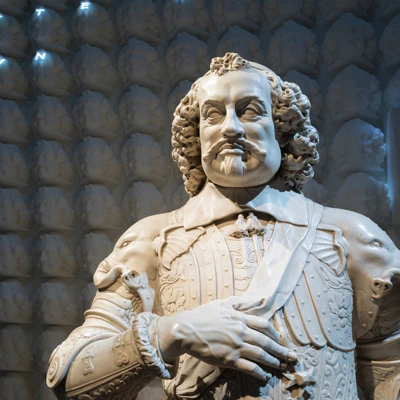 Bust of Johan Maurits in a spotlight, set against a textured wall covered in repeating versions of the same sculpted face. The statue’s elaborate military uniform and expression are clearly visible.