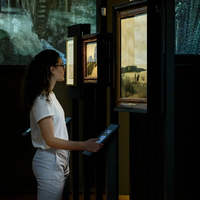 A young woman stands in front of a series of framed landscape paintings, attentively engaging with a touchscreen. Projected scenes of natural environments extend across the walls in the background.