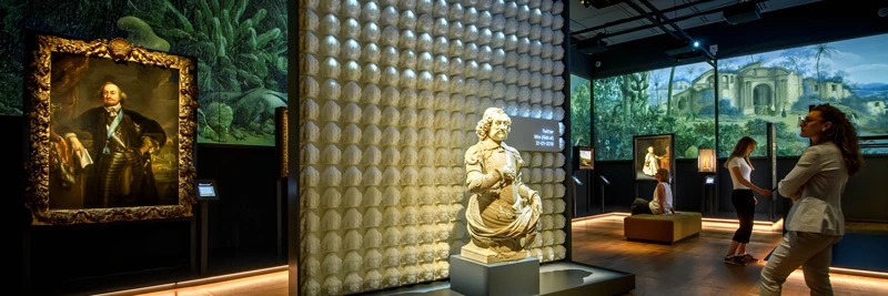 Bust of Johan Maurits positioned in front of a wall covered with identical sculpted heads. Around the room, visitors observe large colonial paintings and immersive wall projections of tropical landscapes and ruins.